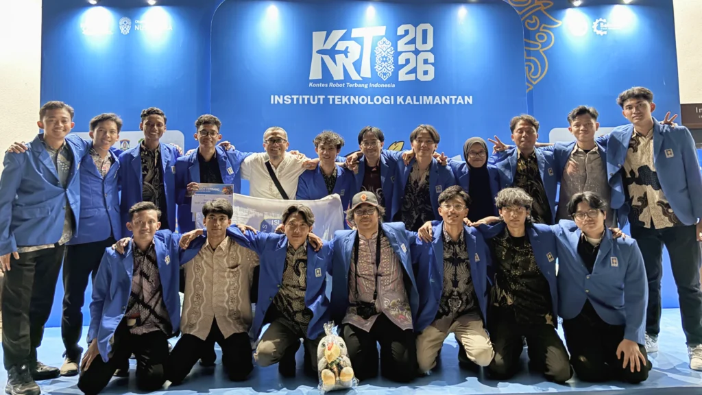 Foto bersama tim besar mahasiswa bersetelan jas almamater biru di atas panggung dengan latar belakang backdrop acara. Latar belakang tersebut berwarna biru terang dengan logo besar "KRTI 2026", teks "Kontes Robot Terbang Indonesia", dan "INSTITUT TEKNOLOGI KALIMANTAN". Beberapa mahasiswa di depan berlutut, sementara yang lain berdiri di belakang, semuanya tersenyum ke arah kamera.