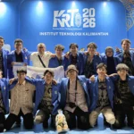 Foto bersama tim besar mahasiswa bersetelan jas almamater biru di atas panggung dengan latar belakang backdrop acara. Latar belakang tersebut berwarna biru terang dengan logo besar "KRTI 2026", teks "Kontes Robot Terbang Indonesia", dan "INSTITUT TEKNOLOGI KALIMANTAN". Beberapa mahasiswa di depan berlutut, sementara yang lain berdiri di belakang, semuanya tersenyum ke arah kamera.