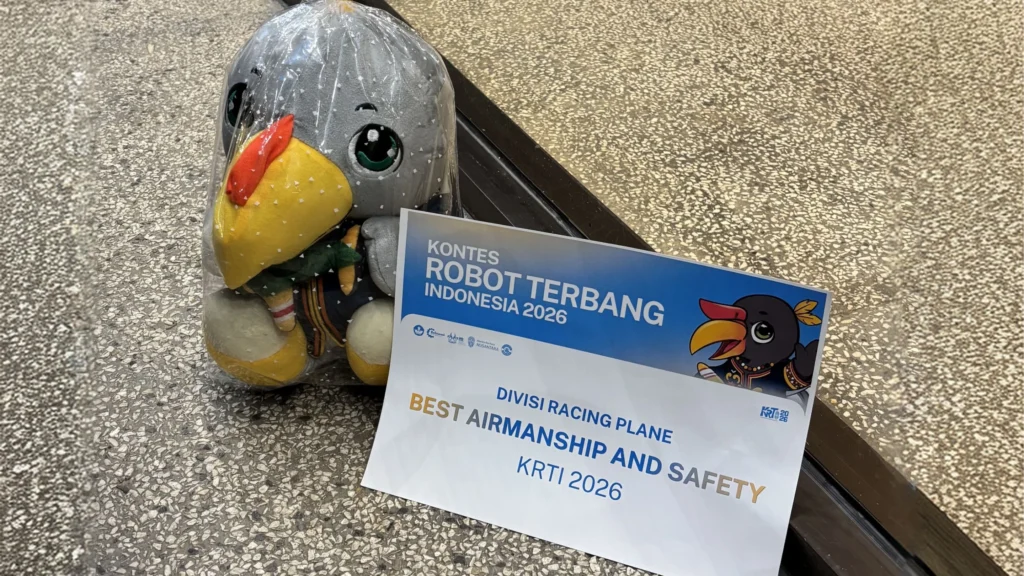 Sebuah boneka maskot burung hantu berwarna abu-abu yang dikemas dalam plastik bening duduk di atas permukaan batu bertekstur kasar. Di sampingnya terdapat sertifikat penghargaan putih dari "KONTES ROBOT TERBANG INDONESIA 2026" yang bertuliskan "DIVISI RACING PLANE" dan "BEST AIRMANSHIP AND SAFETY KRTI 2026", lengkap dengan logo acara dan siluet burung terbang.